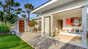 A modern house with a wooden deck, a large glass door leading to a bedroom, and a garden with palm trees. - Villa Momentai (Canggu)