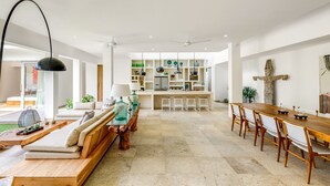 A spacious living area with a sofa, wooden coffee table, and a dining table with chairs. There is a kitchen in the background with a refrigerator and shelves. - Villa Momentai (Canggu)