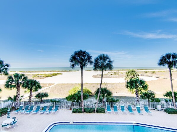 A poolside view with lounge chairs, palm trees, and a beach in the background.