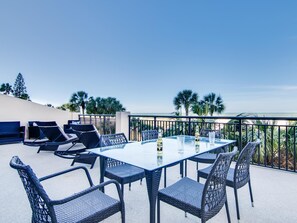A rooftop terrace with a glass table and wicker chairs, overlooking a beach.