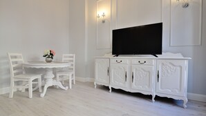Ein Essbereich mit weißem Tisch und Stühlen, einem weißen Schrank mit Flachbildfernseher sowie Wandleuchten.