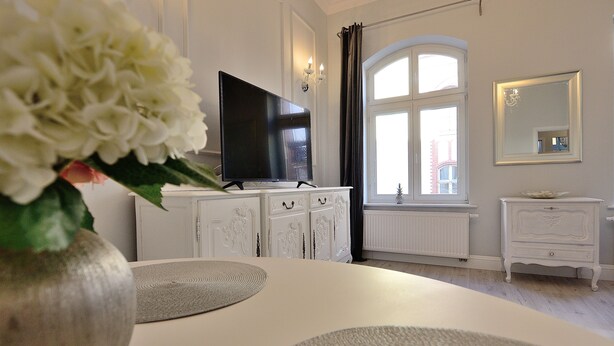 Ein modernes Wohnzimmer mit Flachbildfernseher, weißen Schränken, einem großen Fenster mit Vorhängen und einem Spiegel an der Wand.