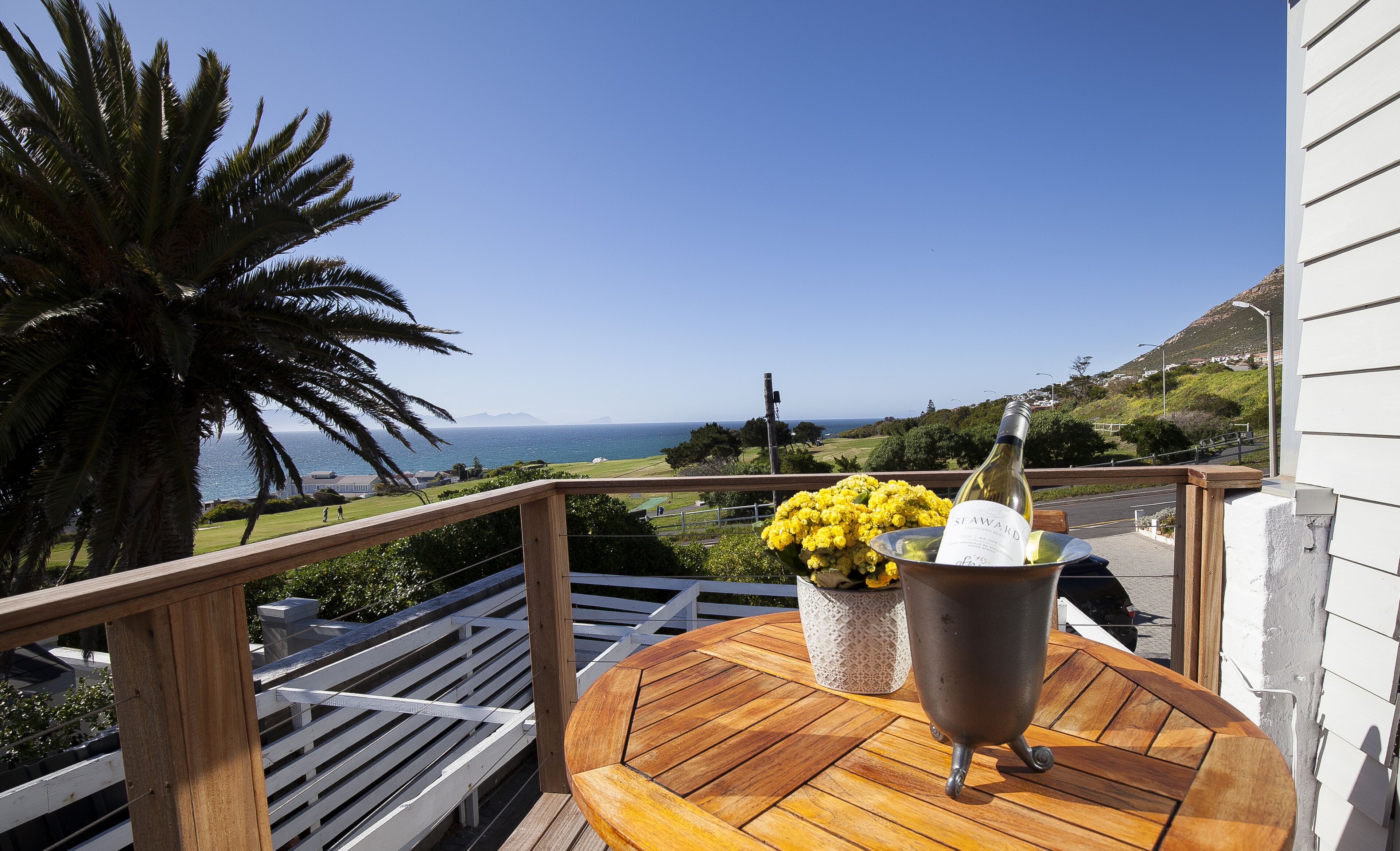 A wooden deck with a table and a metal bucket containing a bottle of wine and yellow flowers.