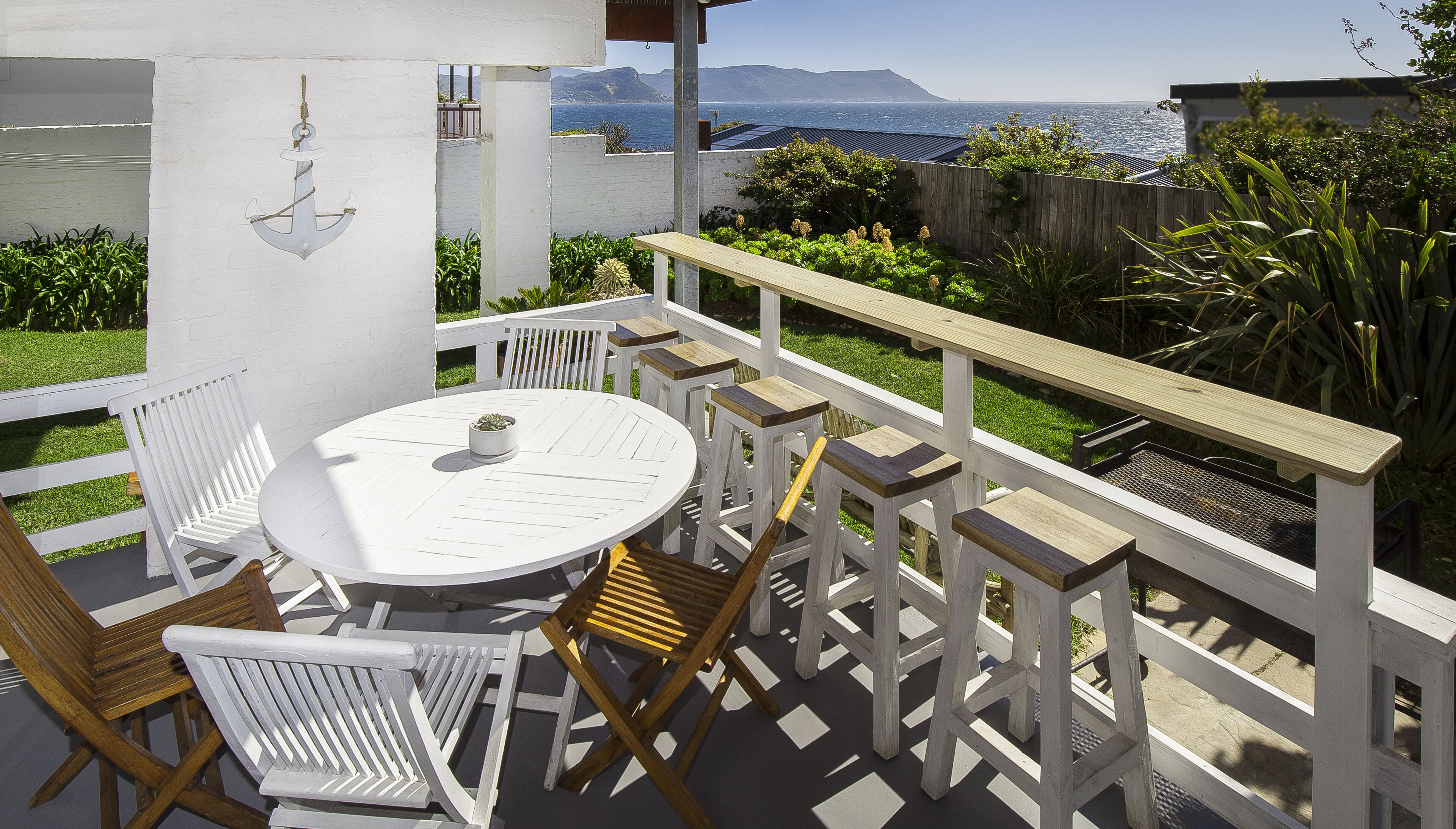 A patio with a round table and white chairs, overlooking a body of water and a mountainous landscape.