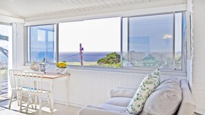 A room with a large window overlooking the sea, a white chair, a small table with a vase of flowers, and a sofa with a cushion. - Penguins View Guesthouse (Cape Town)