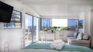 A bedroom with a large window, a bed, a chair, a table with a vase of flowers, and a painting on the wall. - Penguins View Guesthouse (Cape Town)