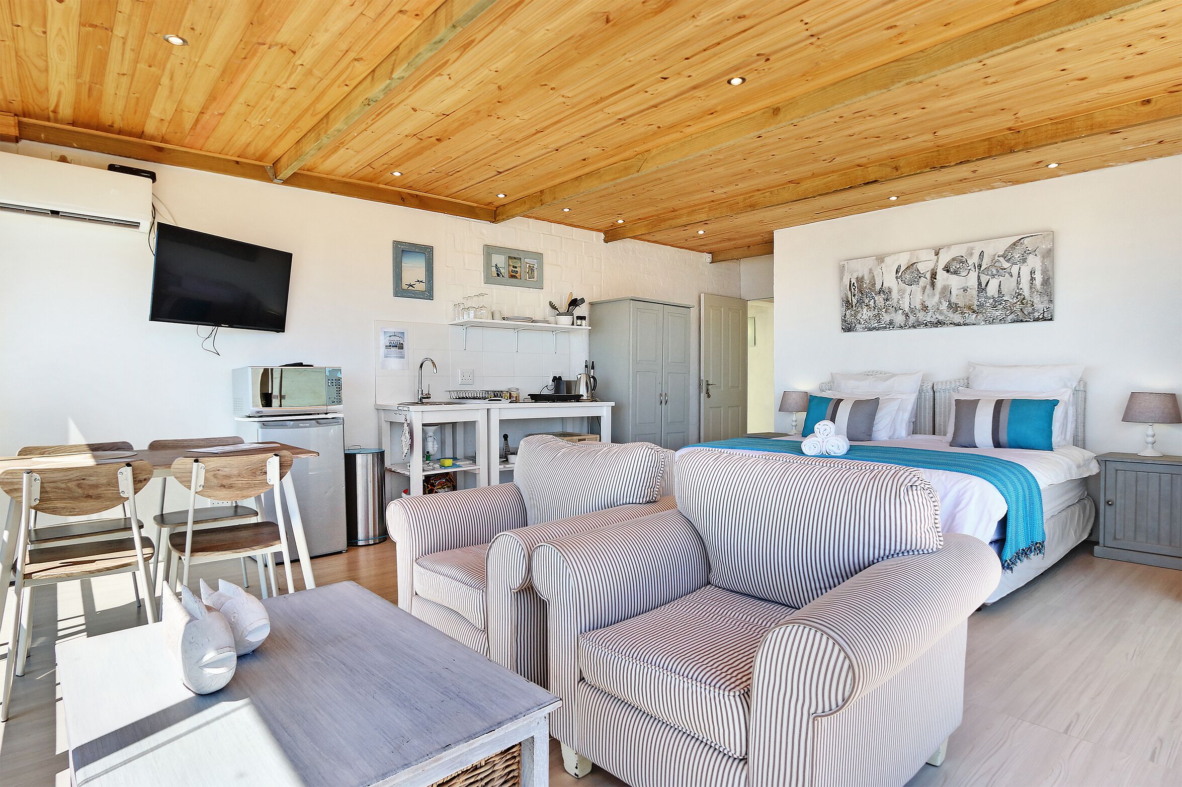 A modern, well-lit room with a wooden ceiling, a bed with striped bedding, a dining area with chairs and a table, and a kitchen area with a TV and cabinets.