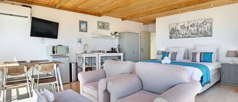 A modern, well-lit room with a wooden ceiling, a bed with striped bedding, a dining area with chairs and a table, and a kitchen area with a TV and cabinets.