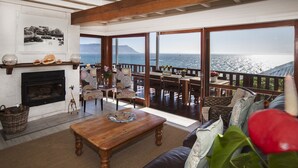 A spacious living area with a fireplace, wooden furniture, and large windows overlooking the sea. - Penguins View Guesthouse (Cape Town)
