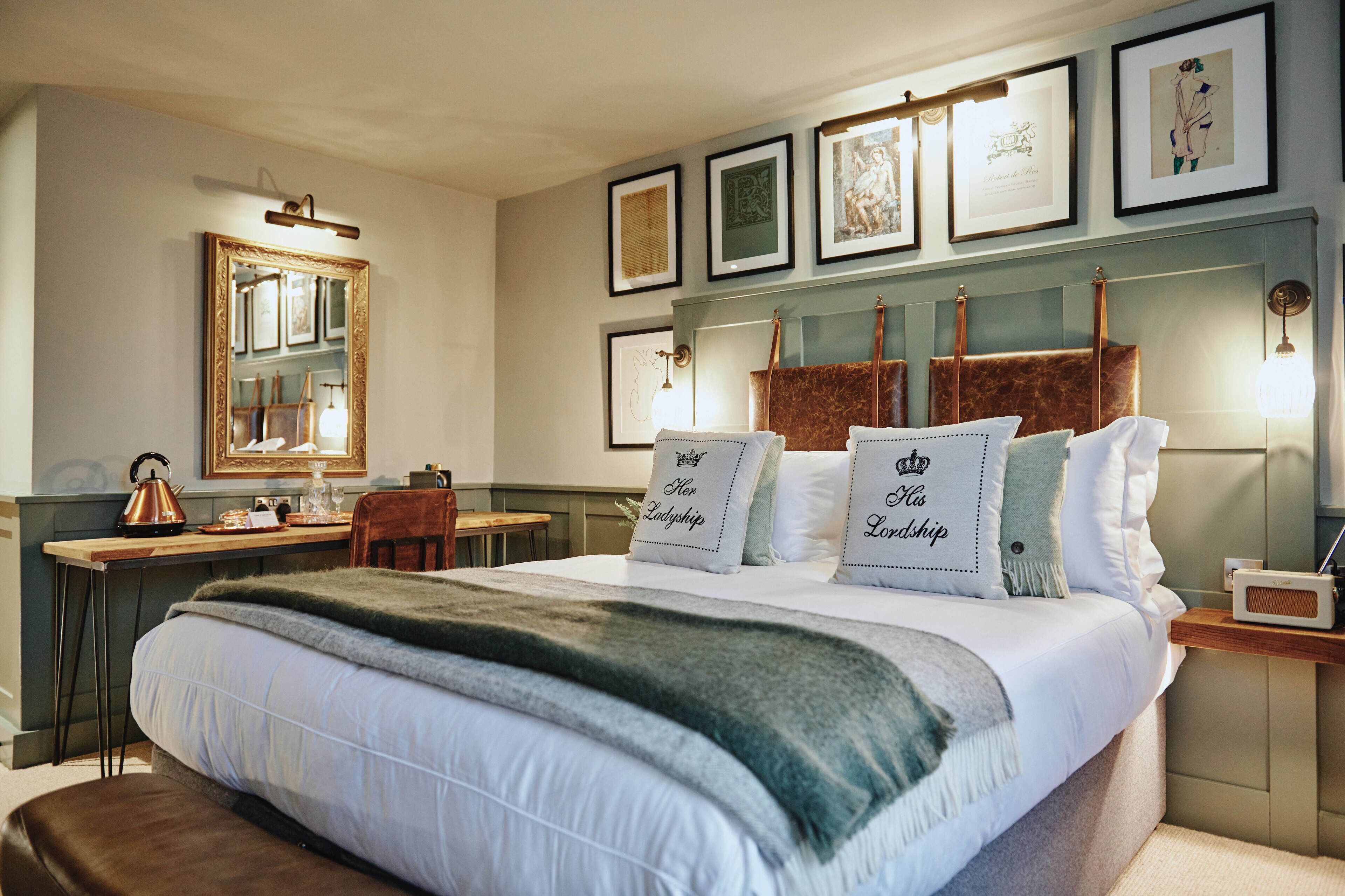 Une chambre bien rangée, avec un grand lit, un bureau et une chaise. Des tableaux encadrés ornent le mur, et un miroir est placé au-dessus du bureau.