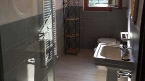 A bathroom with a glass shower enclosure, a white pedestal sink, a window with a view of trees, and a metal shelving unit. - Frantoio-Large and Beautiful family apartment -Agriturismo San Jacopo (Reggello)