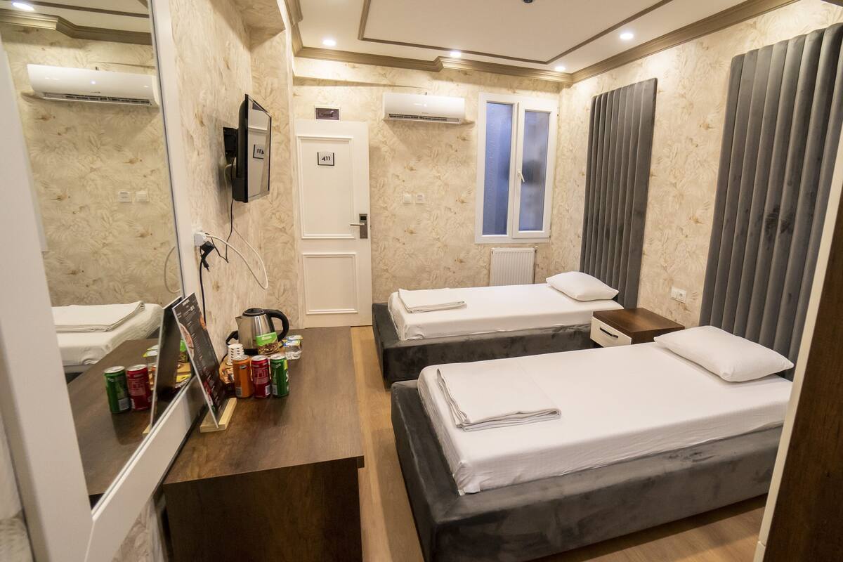 A hotel room with two beds, a small table, a mirror, and a window with curtains.