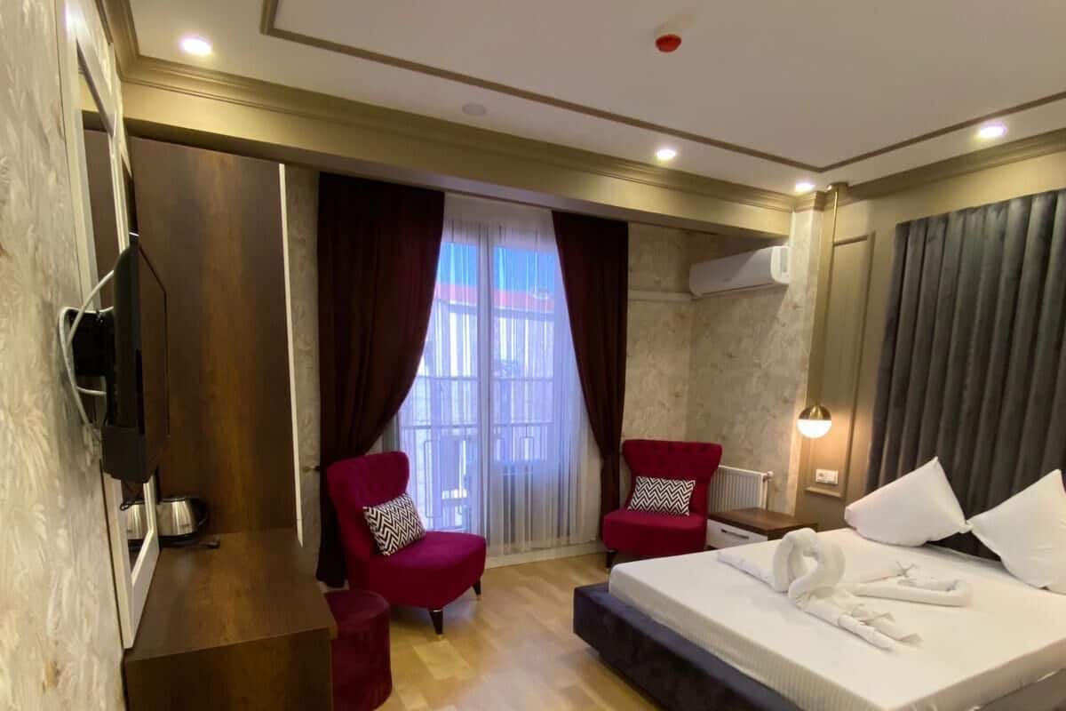 A hotel room with a bed, two red armchairs, a wooden desk, and a large window with curtains.