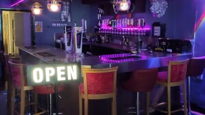 A bar area with a neon sign indicating it's open, bar stools, a counter with a cash register, and a shelf with bottles. - Mardi Gras Hotel (Blackpool)