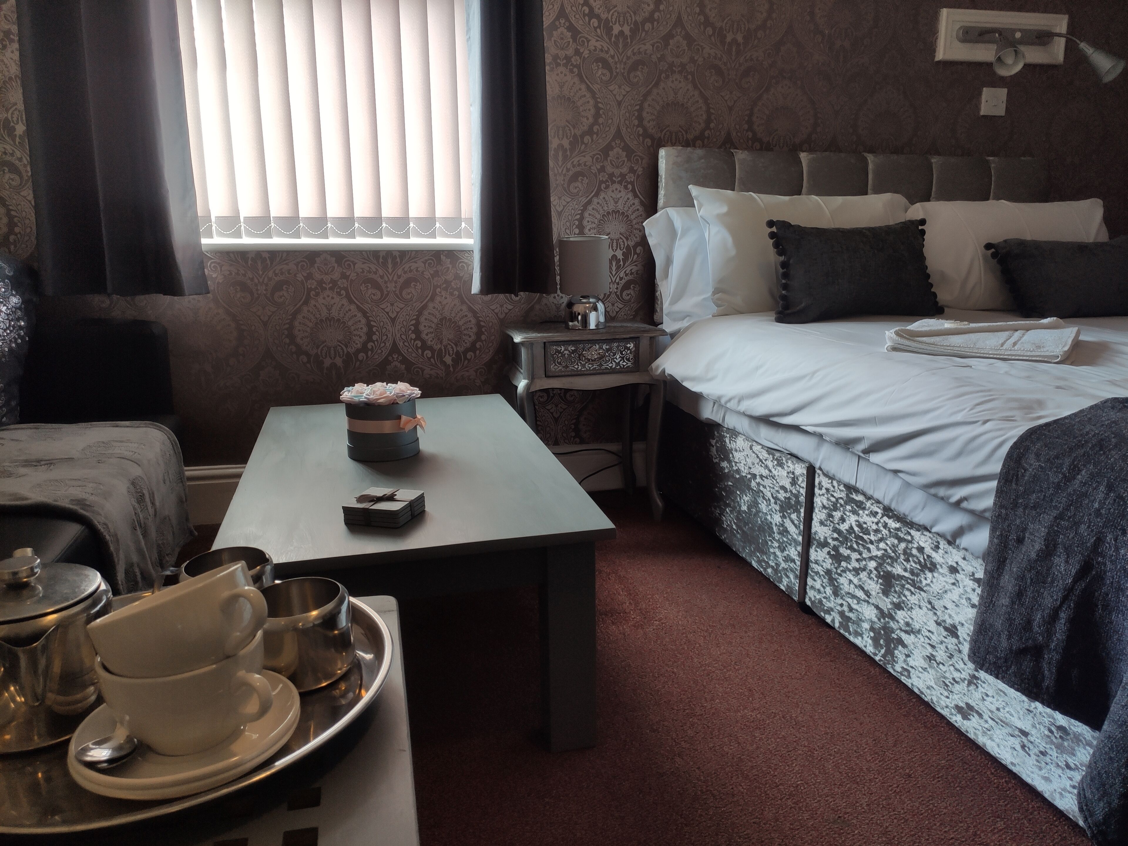 Ein Hotelzimmer mit einem ordentlich bezogenen Bett, einem Nachttisch mit Lampe, einem kleinen Tisch mit einer Kaffeebahn und einem Fenster mit Jalousien.