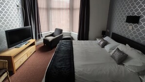 A hotel room with a bed, a TV on a wooden stand, a small seating area with a chair and ottoman, and a window with curtains. - Mardi Gras Hotel (Blackpool)