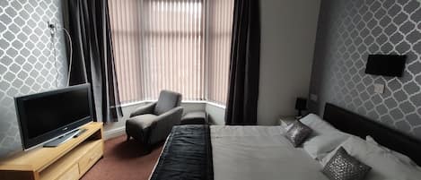 Une chambre d’hôtel avec un lit, un téléviseur posé sur un meuble en bois, un petit coin salon avec un fauteuil et un pouf, et une fenêtre avec des rideaux.