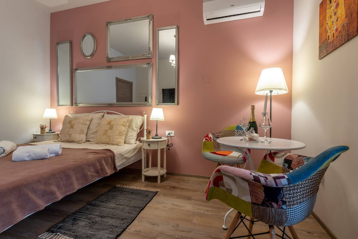 A bedroom with a bed, bedside lamps, a small round table with a lamp, a chair, and a painting on the wall.