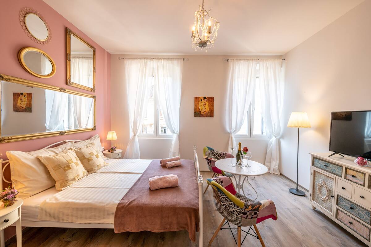 A bedroom with a bed, bedside tables, a TV, a dining table with chairs, and a chandelier.