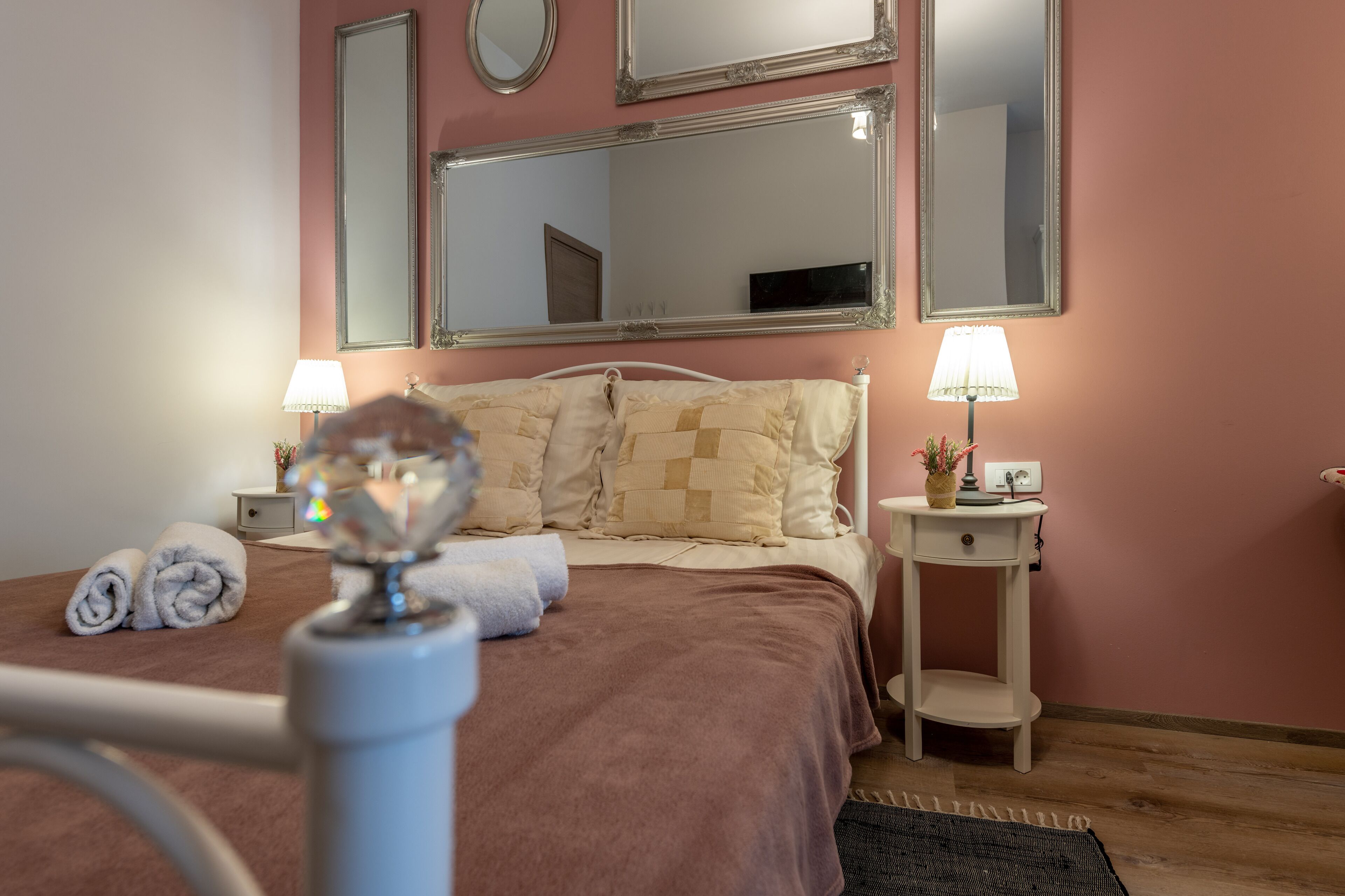 A neatly made bed with a brown blanket, two bedside lamps, a small bedside table with a lamp, and a mirror above the bed.