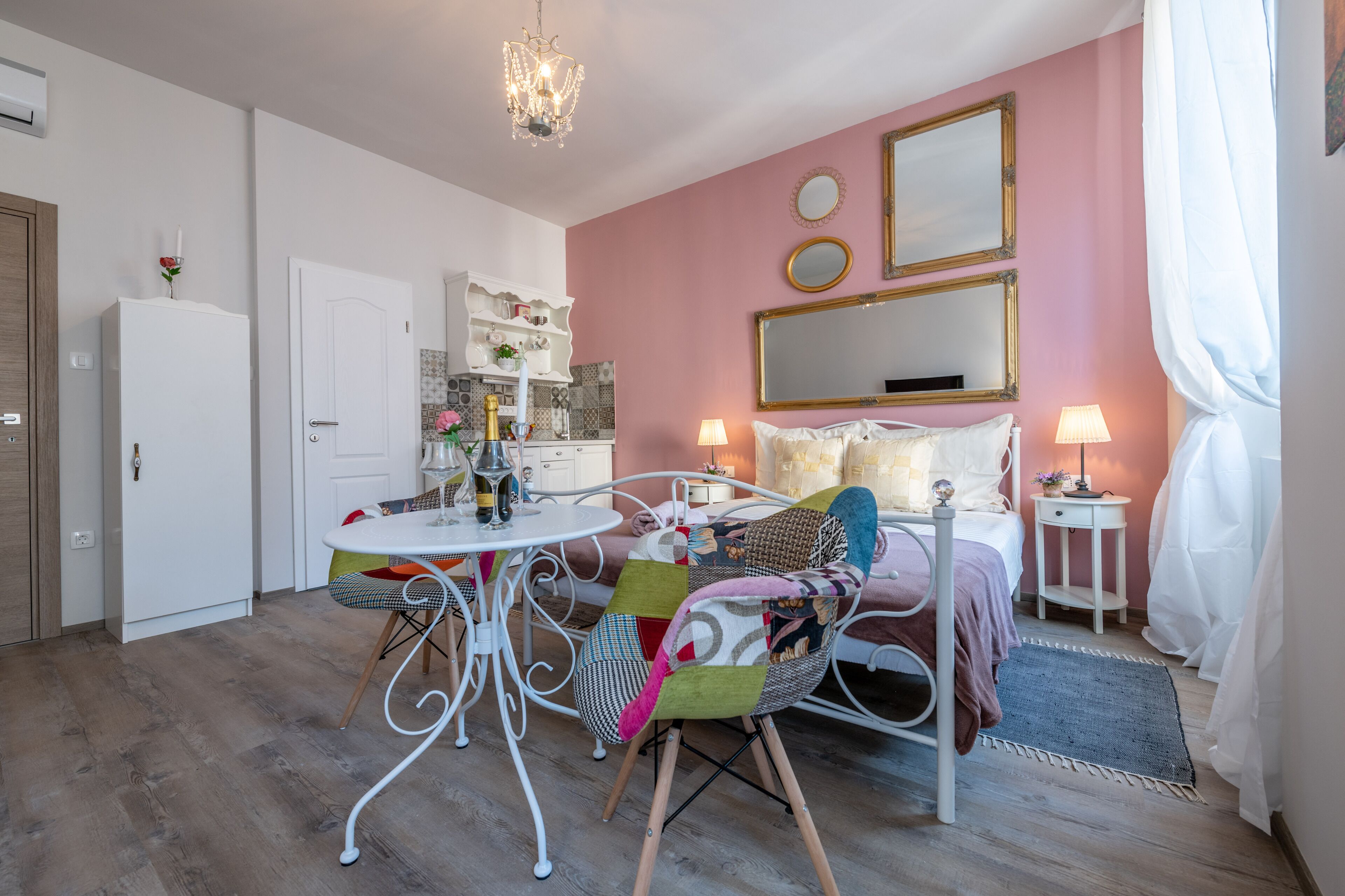 A bedroom with a bed, bedside tables, a nightstand, a window with curtains, a chandelier, and a small sitting area with a table and chairs.
