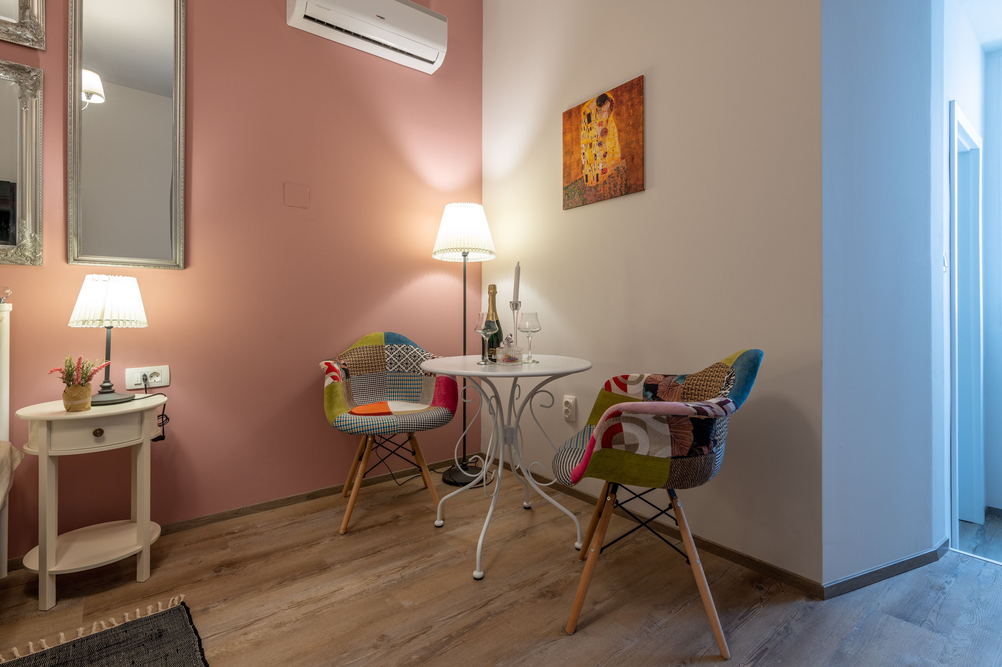 A room with pink walls, a white air conditioner, a table with a lamp, two chairs, and a small white side table with a lamp.