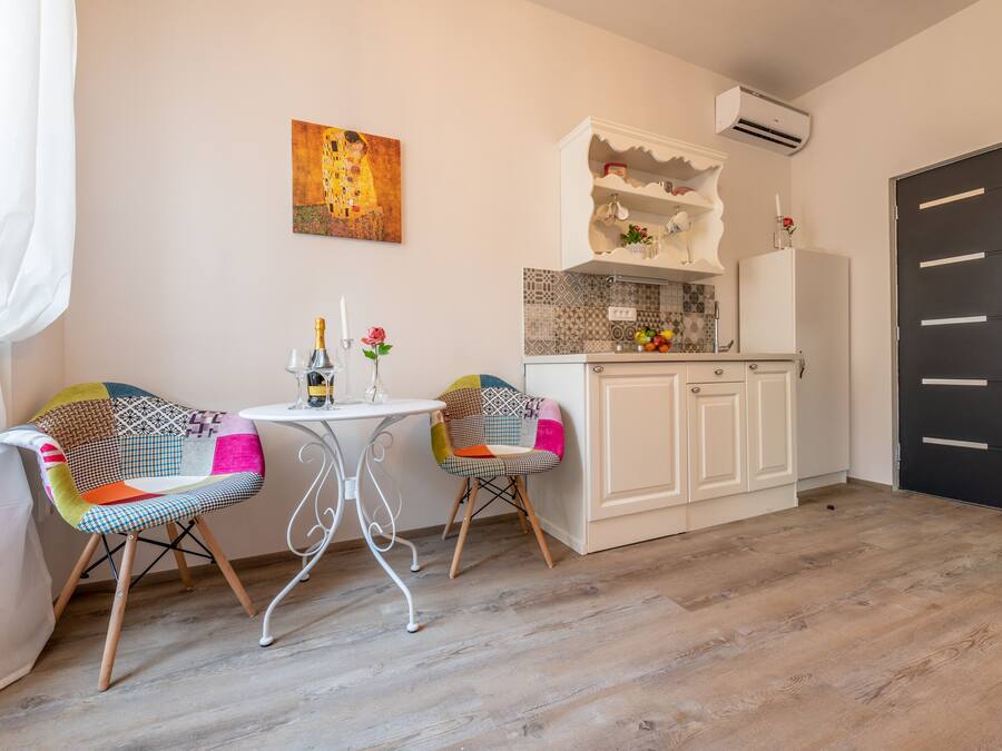 A compact kitchen with white cabinets, a tiled backsplash, and a black door. There is a small table with two chairs, one with a colorful cushion. A painting hangs on the wall above the table.