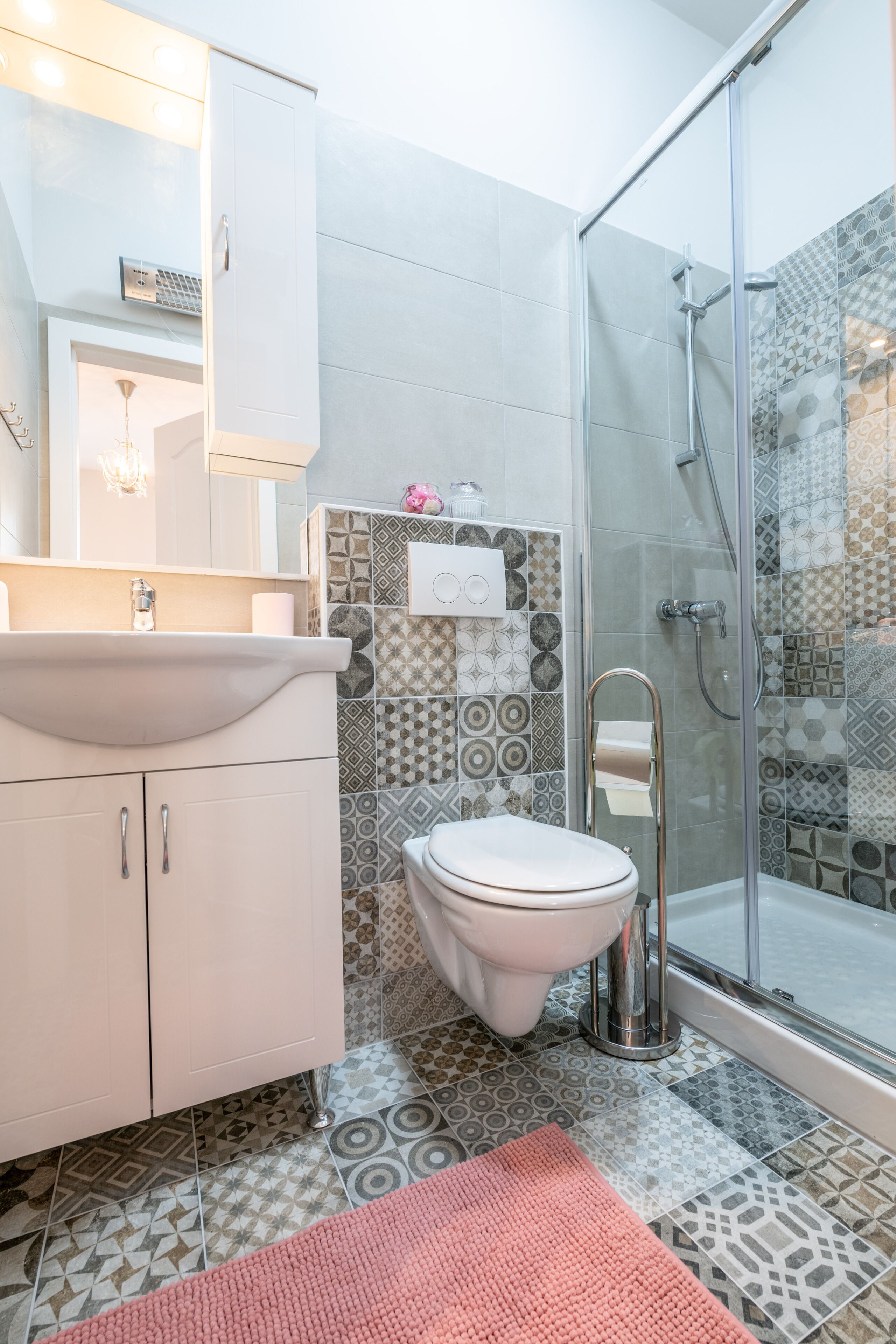 Ein Badezimmer mit einem weißen Waschbecken, einer Toilette und einer Dusche. Der Boden ist mit gemusterten Fliesen ausgelegt, und es liegt ein rosa Teppich vor.