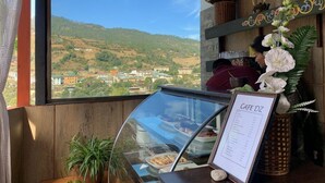 A cafe with a menu board, a glass display case with pastries, a view of a hillside, and wooden decor. - Hotel River Valley (Punakha)