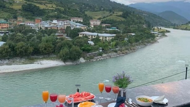 A dining table set with various dishes and drinks overlooking a river and mountains. - Hotel River Valley (Punakha)