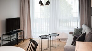 Een moderne woonkamer met een flatscreen televisie, twee kleine tafels, een bank en een groot raam met gordijnen.