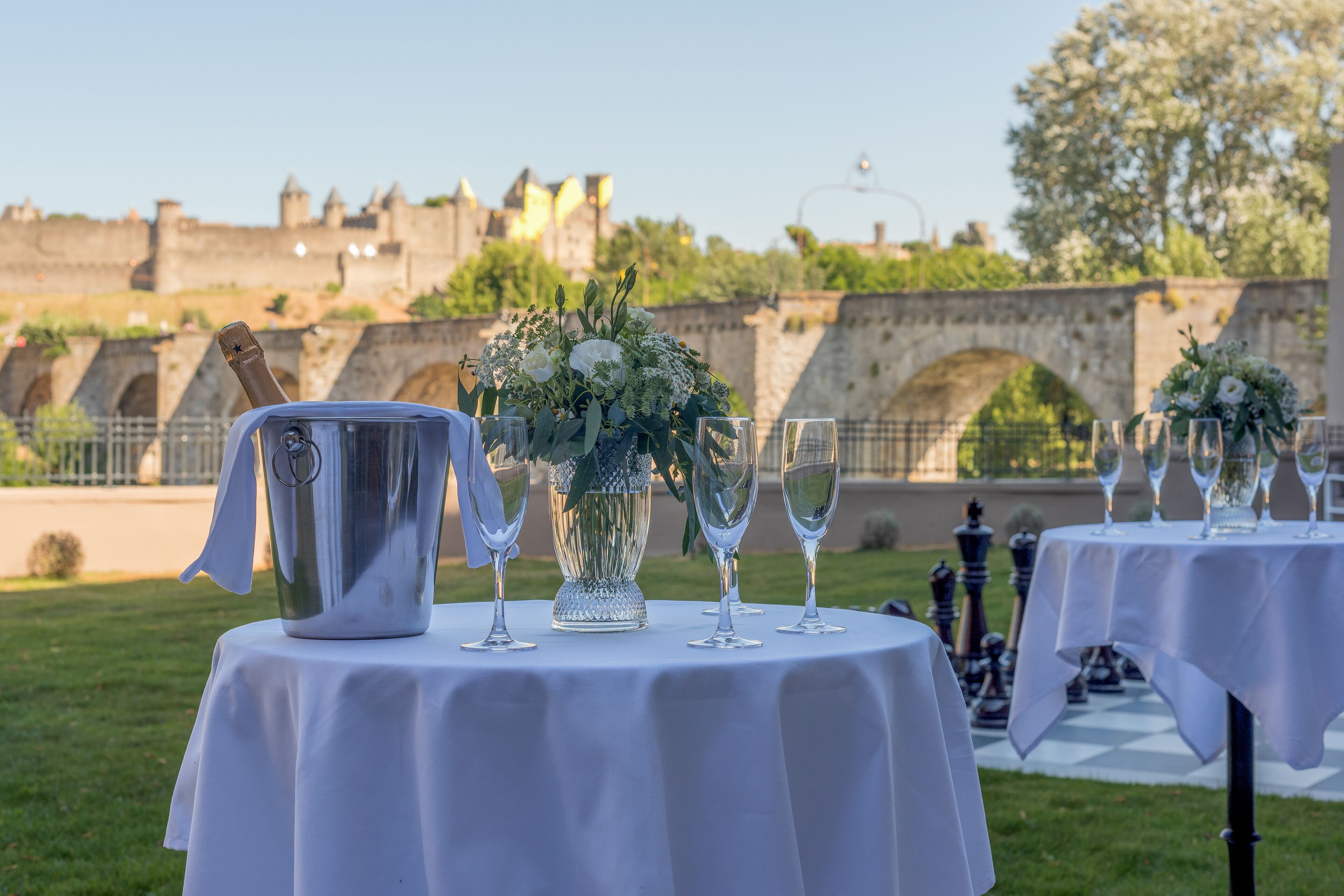 Une table dressée à l’extérieur, avec un pichet en métal, un vase rempli de fleurs et des verres à vin, et en arrière-plan, un pont de pierre historique et un château.