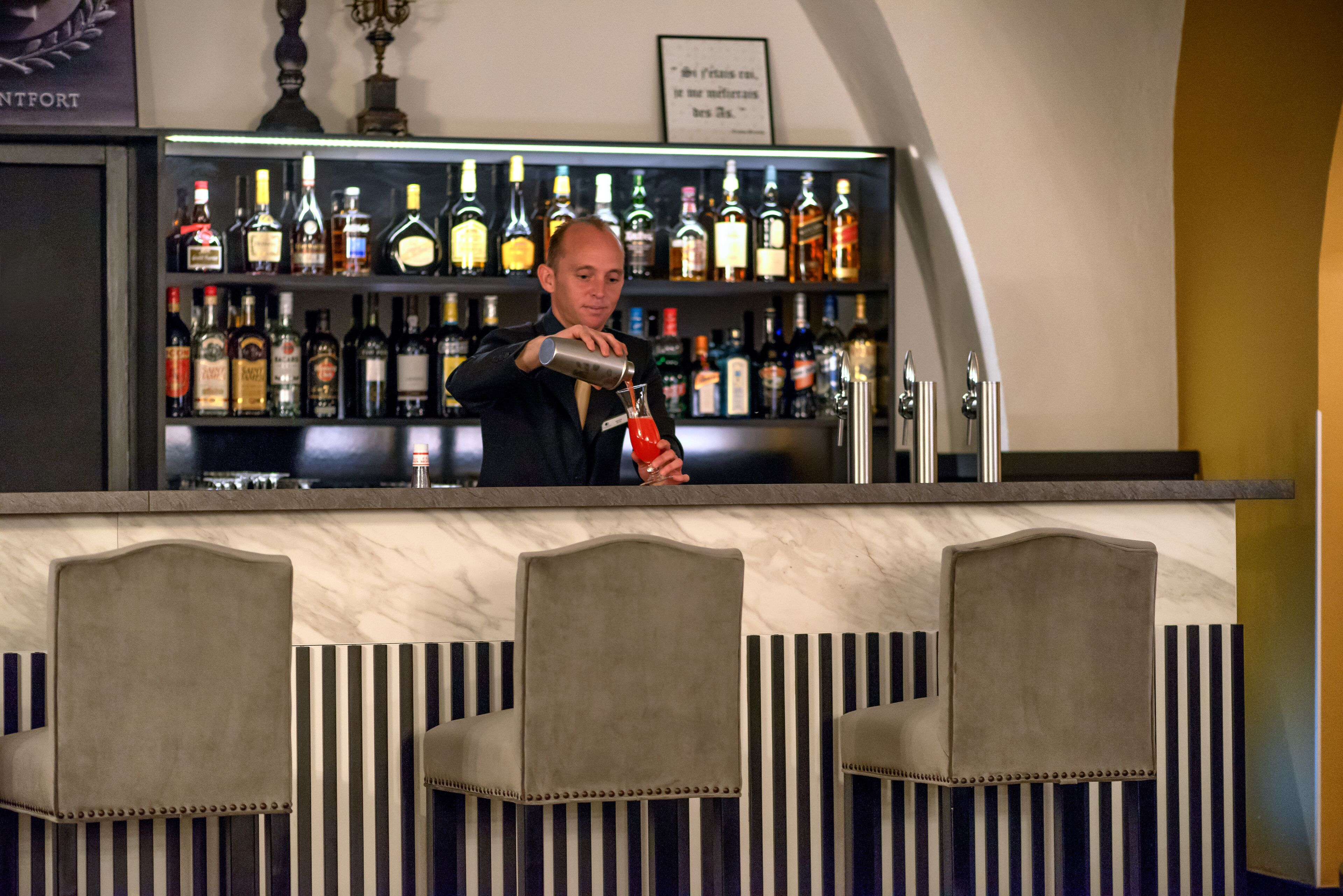 Un espace bar où un barman prépare une boisson. On peut voir des étagères remplies de différentes bouteilles derrière le bar.