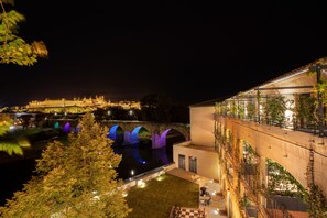 A night view of a historic building with illuminated arches over a river, a modern extension with outdoor seating, and a well-maintained garden area. - SOWELL COLLECTION Hôtel du Roi & SPA (Carcassonne)