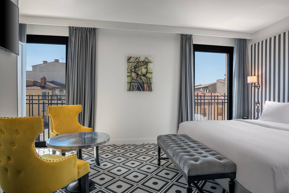A hotel room with a bed, a bench, a round table with two chairs, and a large window with curtains.