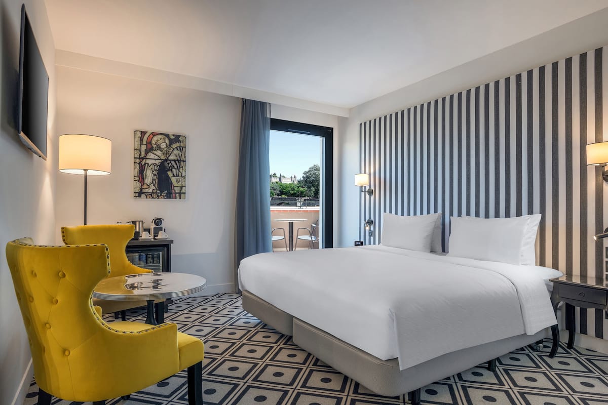 A hotel room with a large bed, a yellow armchair, a small round table, and a view of the outdoors.