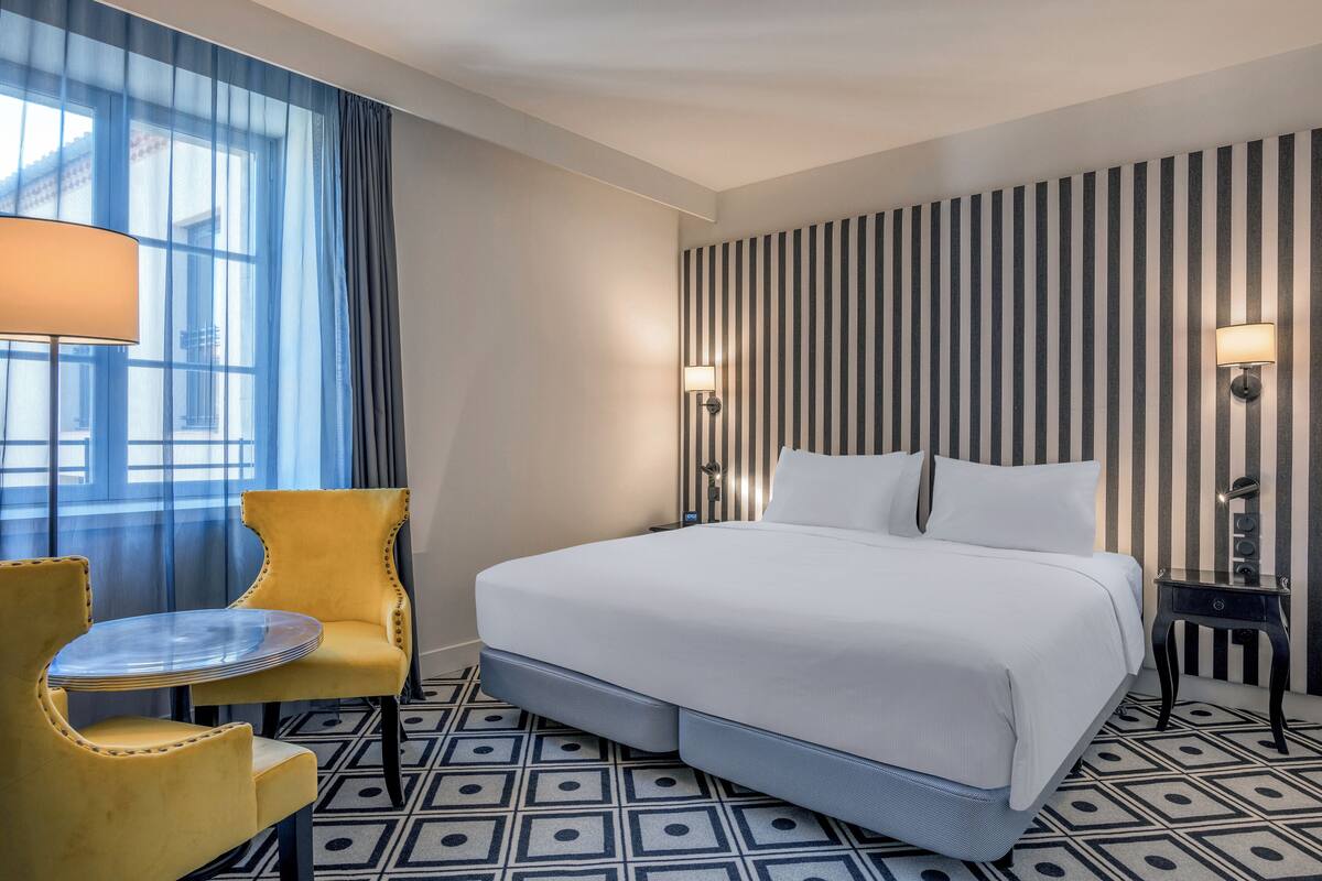 A hotel room with a bed, a chair, a table, and a lamp.