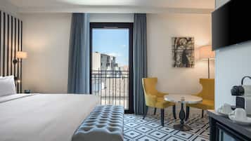 Une chambre d’hôtel comprenant un lit, une banquette, deux chaises, une petite table, un téléviseur et une fenêtre donnant sur des immeubles.