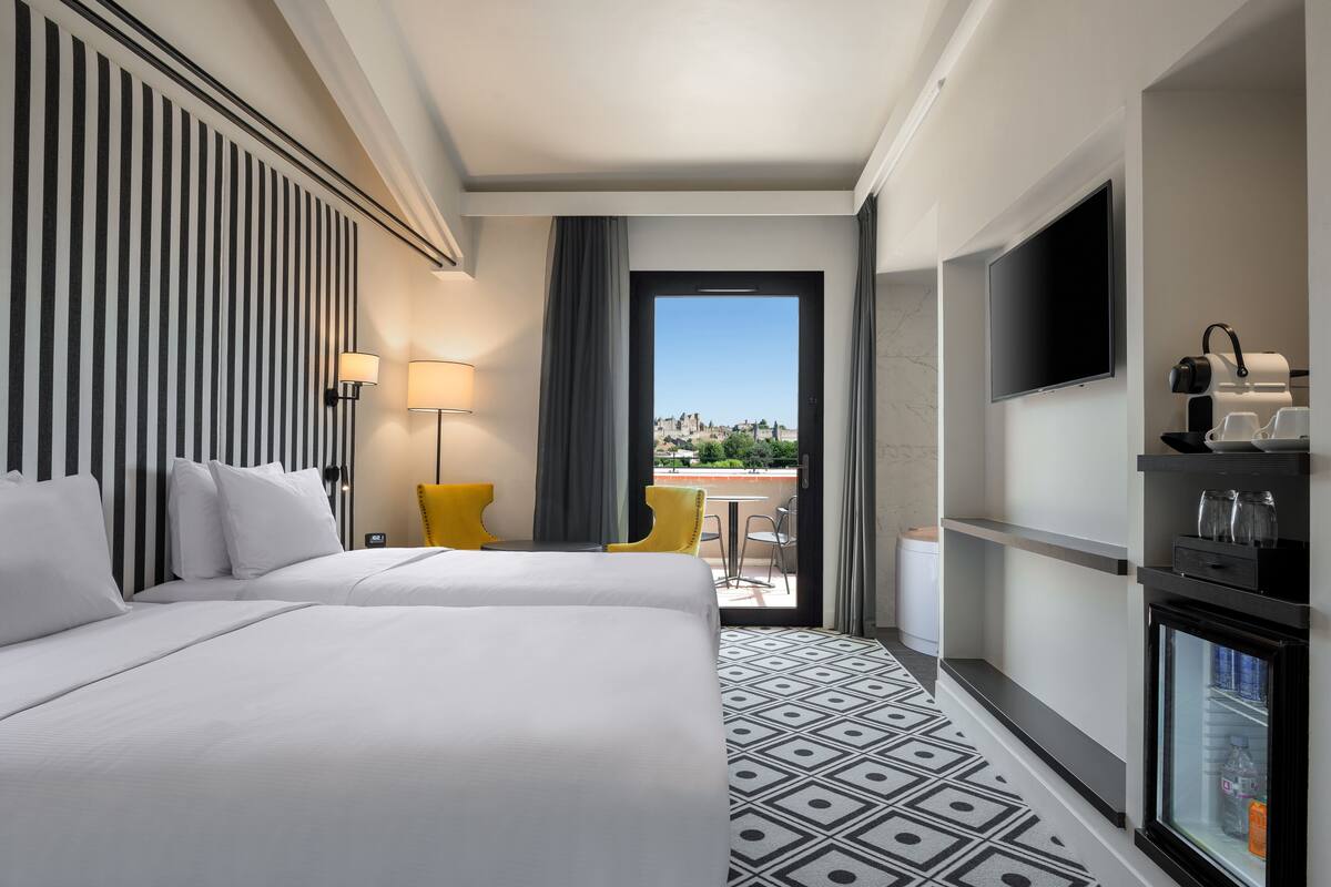 A hotel room with two beds, a TV, a minibar, and a balcony with a city view.