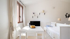 Un dormitorio con cama, mesita de noche y un pequeño escritorio con silla. Hay una ventana con cortinas, un estante en la pared con objetos decorativos y un aplique de pared.