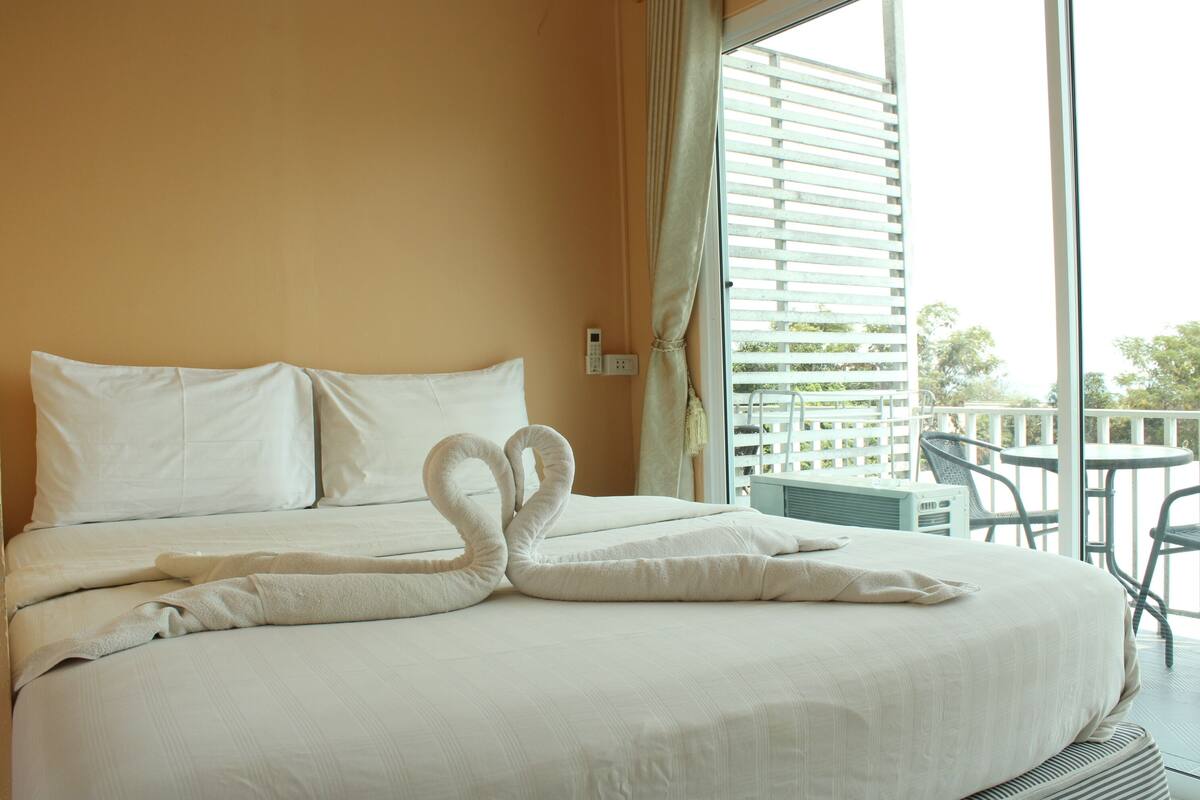 A hotel room with a bed featuring a swan-shaped towel arrangement, a balcony with outdoor furniture, and a view of greenery outside.