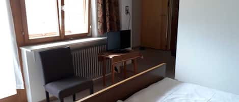 Una habitación con cama, un escritorio con computadora, una silla y un ventanal con cortinas.