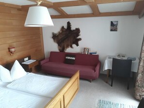 A room with a bed, a red sofa, a wooden headboard, a table with a lamp, and a wall-mounted light fixture. - Double room (type A) Z-NR. 1 - Greber Christine - bed and breakfast Fernblick (Schoppernau)