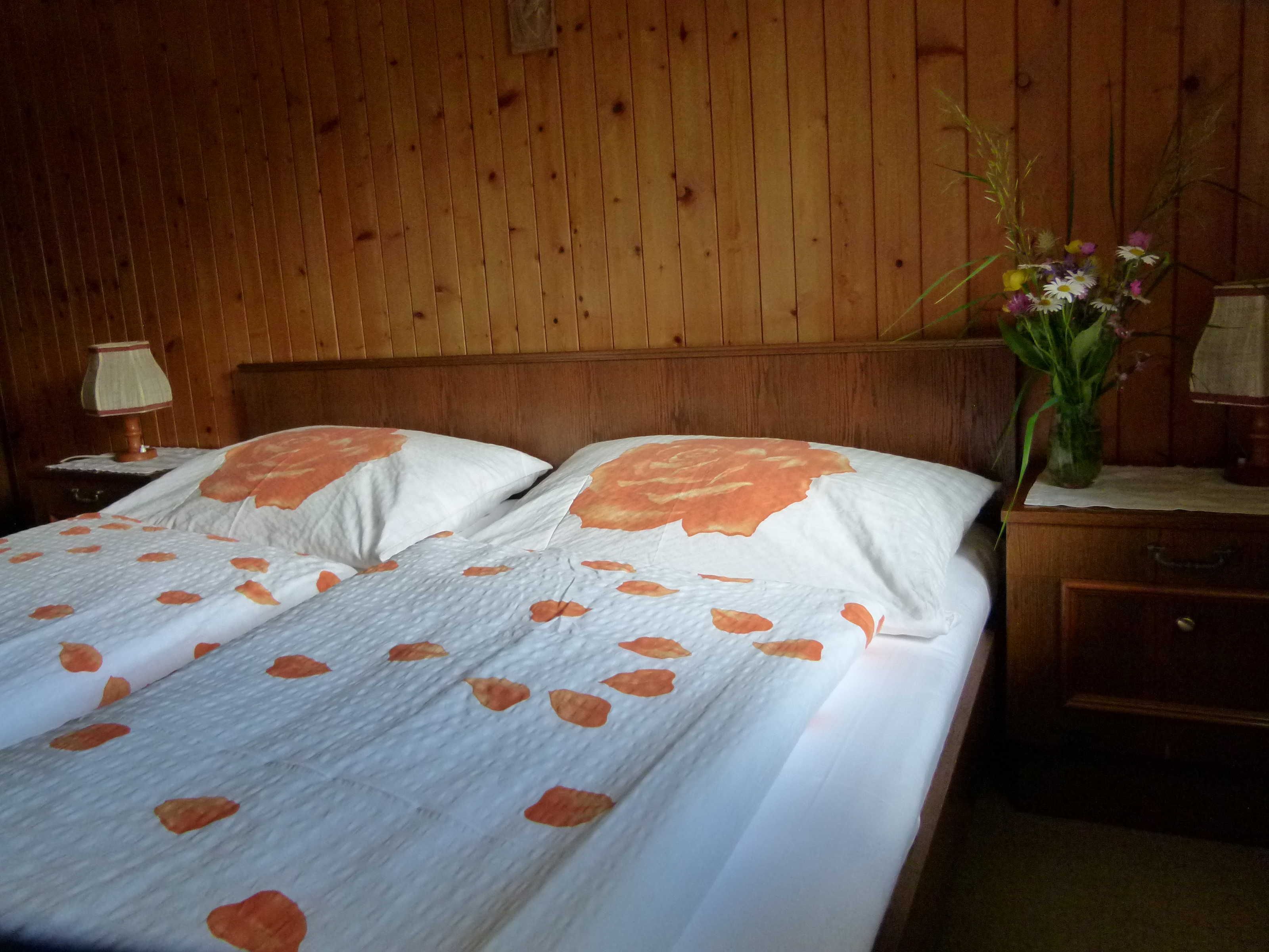 Een kamer met houten lambrisering, een bed, een nachtkastje, een lamp en een vaas met bloemen.