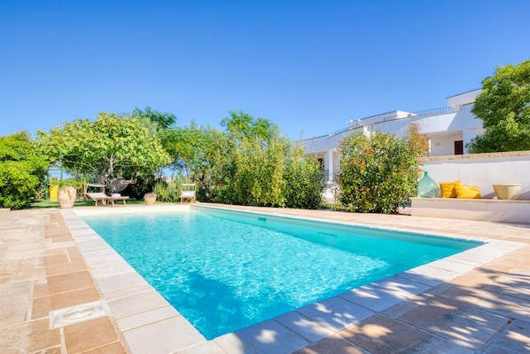 Une piscine dotée d’une terrasse carrelée, entourée de verdure et bordée d’un bâtiment blanc avec un balcon.