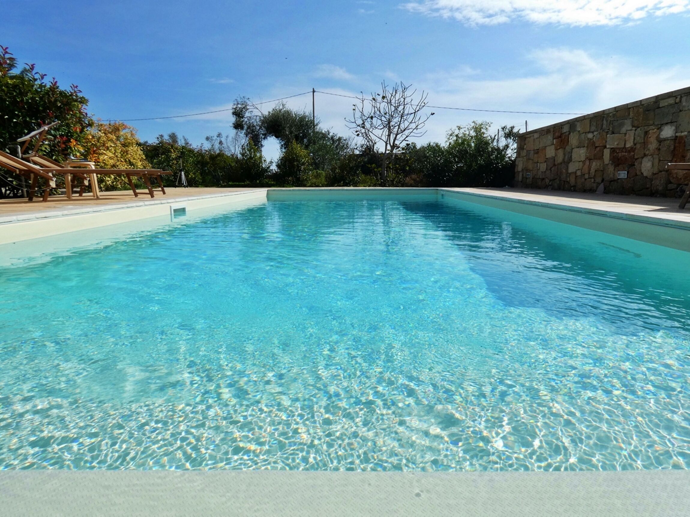 A clear blue swimming pool with a stone wall and a wooden deck on one side.