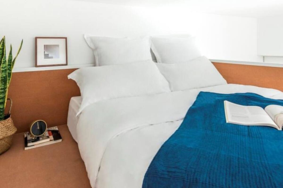 A neatly made bed with white bedding and a blue throw blanket, a small bedside table with a book and a clock, and a framed picture on the wall.
