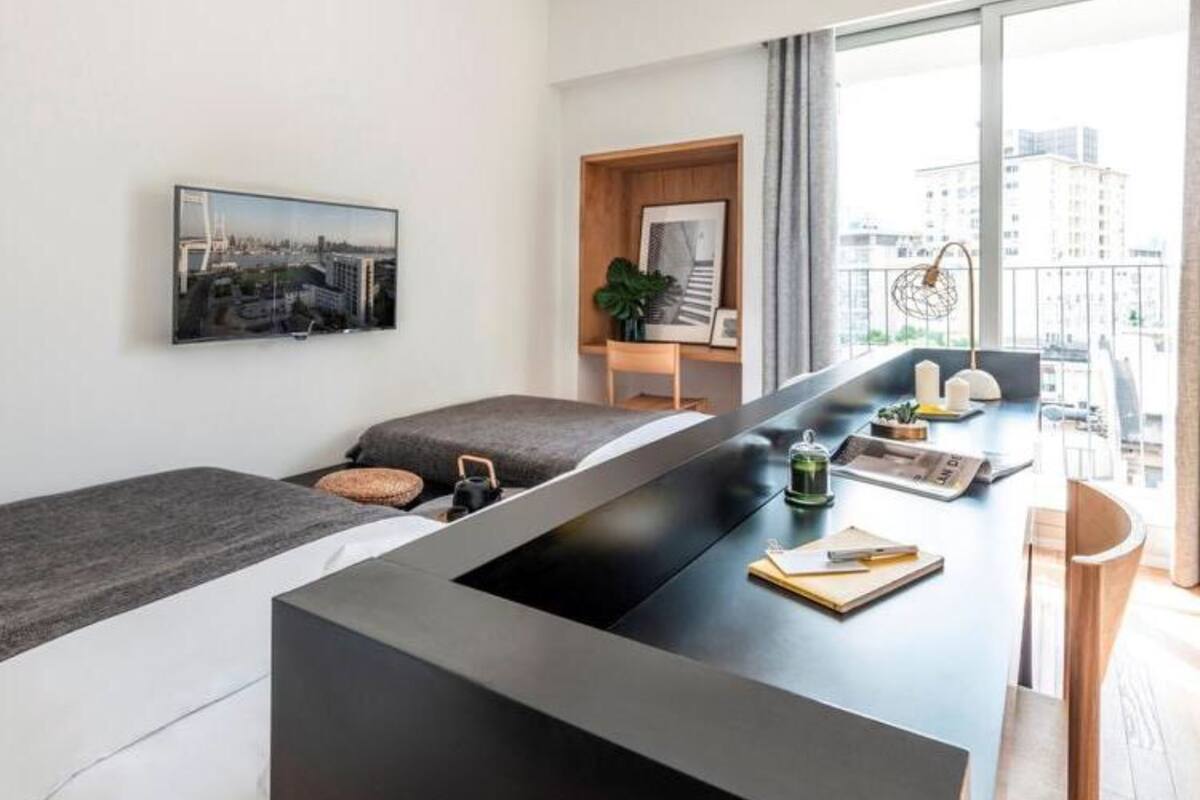 A modern, minimalist room with a central black table, a bed with grey bedding, and a large window offering a city view.