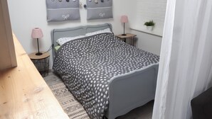 A bedroom with a bed, a bedside table, a lamp, a small table, and a wall-mounted shelf with decorative items. - Independent studio on the 1st floor less than 1 kilometer from the Château de Lavaux (Rochefort)