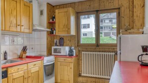 A compact kitchen with wooden cabinets, a red countertop, a microwave, a stove, and a window with a view of a house.